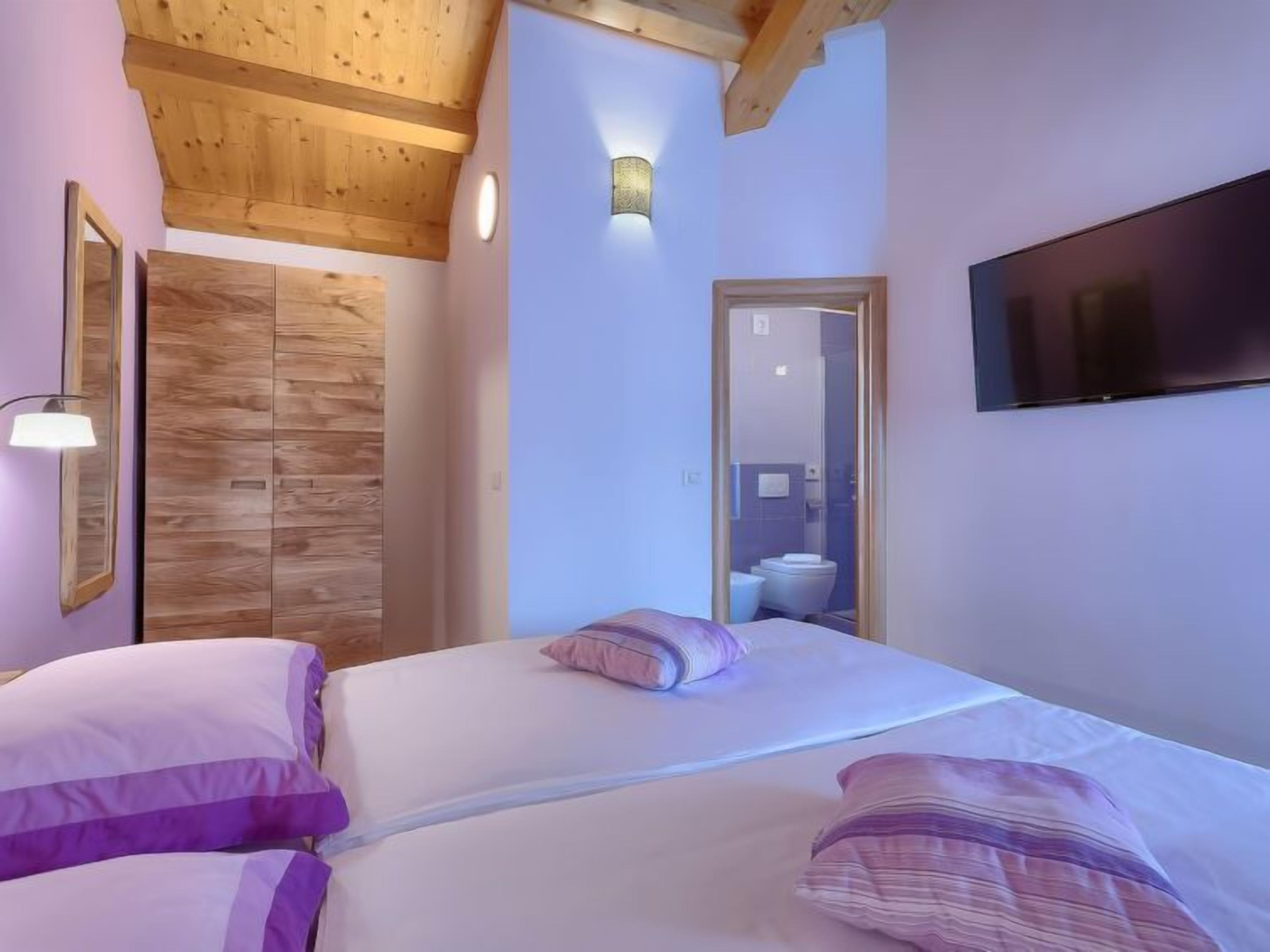 Une chambre avec un plafond en bois, un lit avec une literie blanche et violette, un téléviseur fixé au mur et une armoire en bois.