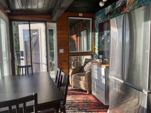 A kitchen with a stainless steel refrigerator, wooden cabinets, a dining table with chairs, and a large window with a view of the outdoors.