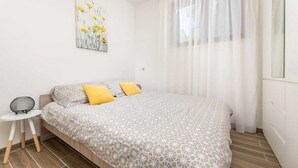 A bedroom with a bed, a small side table, a wall-mounted artwork, and a window with sheer curtains. - Apartment LaPePa (Silo - island Krk)