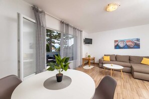 Uma sala de estar moderna com um sofá, uma mesa de centro, uma TV fixada na parede e uma grande janela com cortinas.