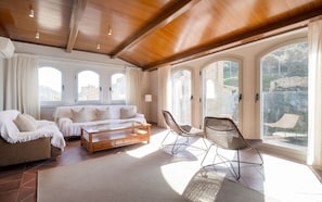 A spacious living area with a wooden ceiling, a sofa, two chairs, a coffee table, and large windows offering a view of the outdoors. - Luxury Rocamar – Beachfront & Terrace (Tossa De Mar)