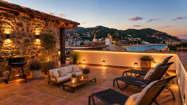 A rooftop terrace with stone walls, a sofa, lounge chairs, and a view of a coastal town at dusk. - Luxury Rocamar – Beachfront & Terrace (Tossa De Mar)