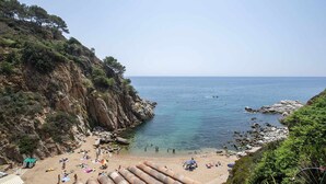 A coastal beach with rocky cliffs, clear blue sea, and people sunbathing. - Luxury Rocamar – Beachfront & Terrace (Tossa De Mar)