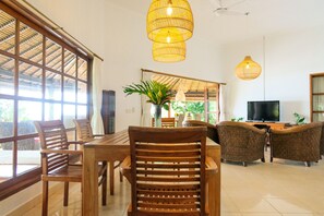 A spacious living room with a wooden dining table, two sofas, and a TV. Large windows allow natural light to fill the room. - Villa Cahaya, beachfront, 2 Bedrooms w/ensuite baths, private pool fully staffed (lokapaksa)