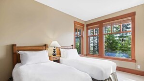 Habitación de hotel con dos camas, una mesita de noche con lámpara y ventanas amplias con vista a árboles.
