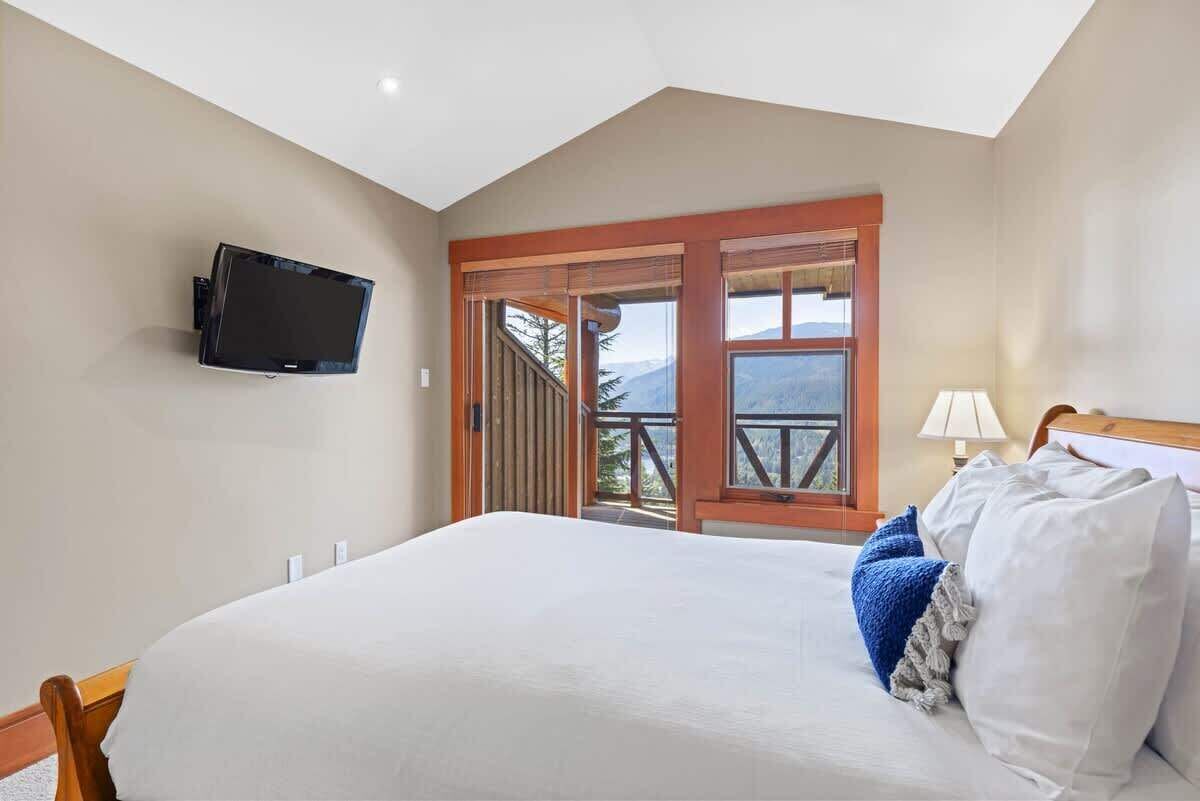 A bedroom with a bed, a mounted TV, a window with a view of trees and mountains, and a wooden door.