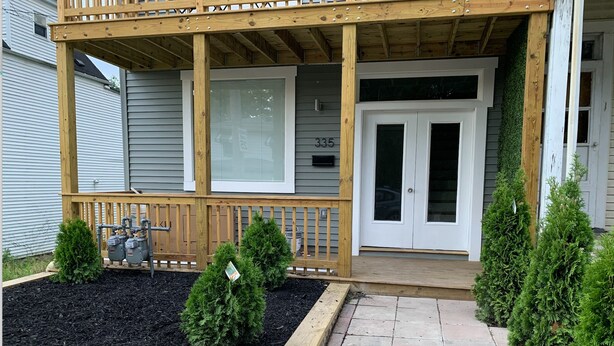 A covered porch with a wooden railing, a stone pathway, and a small garden area with mulch and young trees. - New 2 Bed 2Bath Spacious Apartment w/Deck & Parking (Pittsburgh)