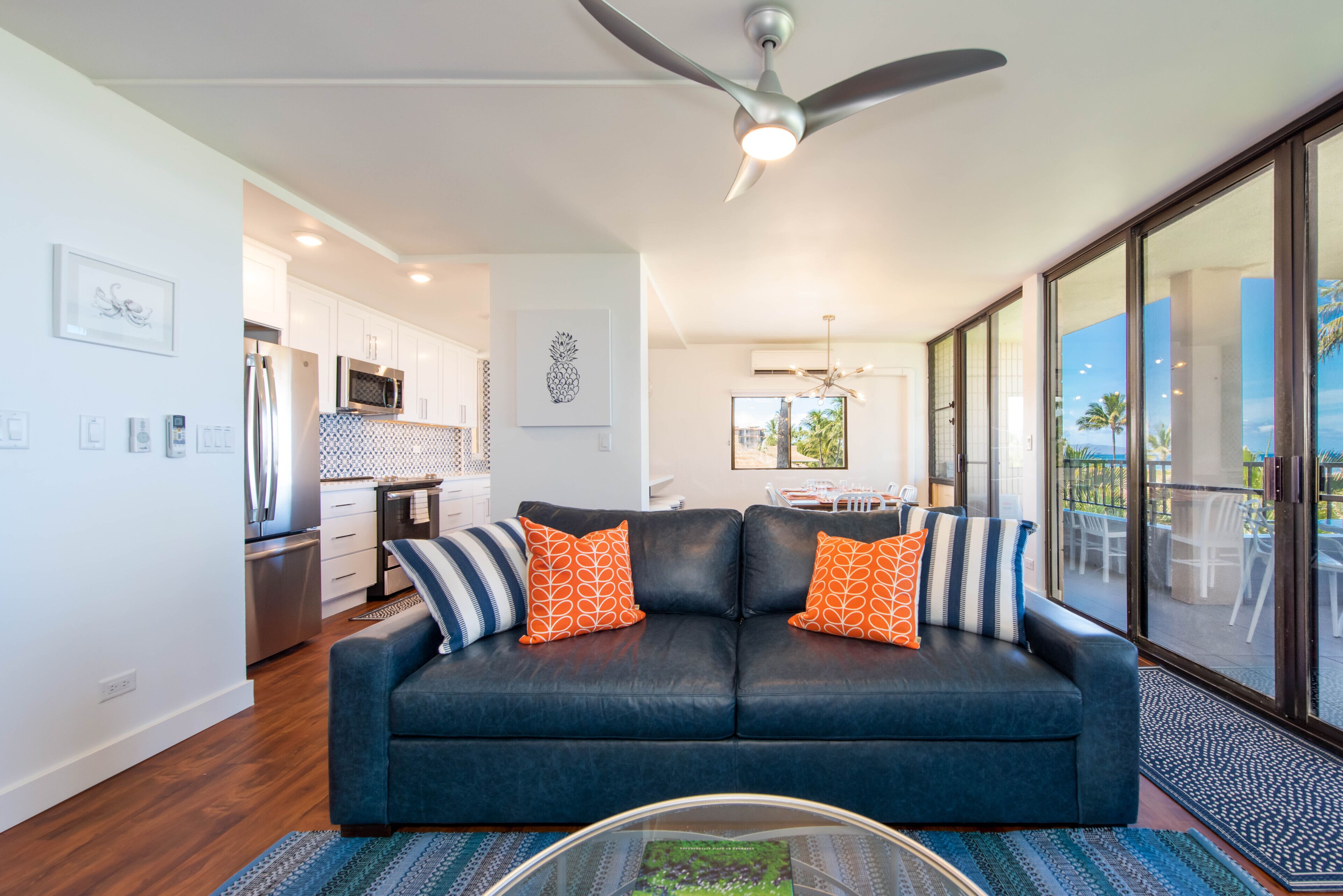 A modern living room with a dark blue sofa, two orange patterned pillows, a glass coffee table, and a ceiling fan.