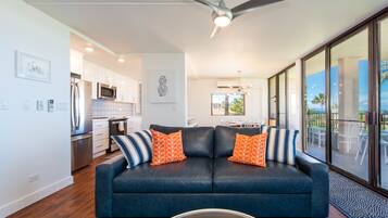 A modern living room with a dark blue sofa, two orange patterned pillows, a glass coffee table, and a ceiling fan.