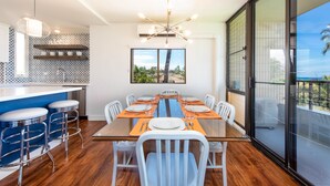 Un comedor moderno con una mesa de vidrio, sillas blancas y manteles individuales anaranjados. Se observan taburetes y una cocina al fondo.