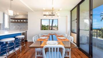 A modern dining area with a glass table, white chairs, and orange placemats. There are bar stools and a kitchen in the background.