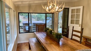 Ein Essbereich mit Holztisch, Bänken, einer Chandelüster und Blick ins Freie durch große Fenster.