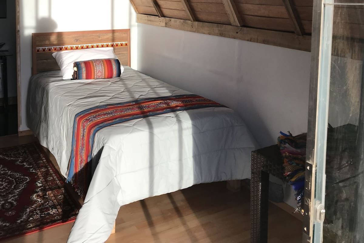Un dormitorio con techo inclinado, una cama con colcha estampada, cabecera con diseño geométrico y ventana con vista a un edificio.