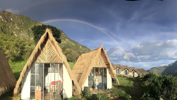 A row of thatched-roof cottages with glass doors, set against a mountainous backdrop with a rainbow. - Choquequirao Sanctuary Lodge (Santa Teresa)