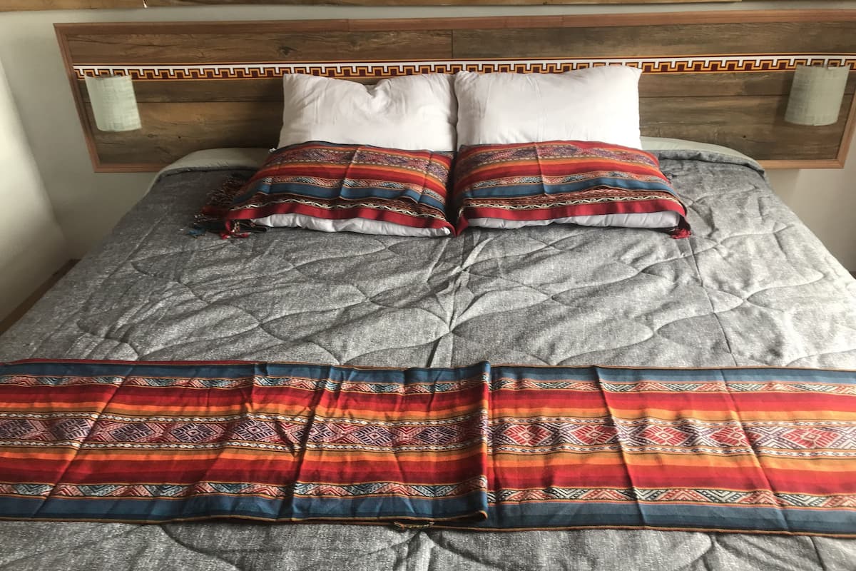 Una cama con edredón gris acolchado, dos almohadas blancas y dos mantas coloridas con diseños geométricos.