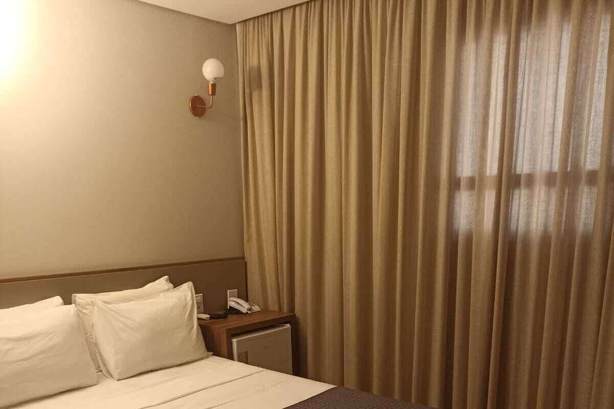 Ein Hotelzimmer mit einem großen Bett, einem Nachttisch mit Lampe und einem Fenster mit Vorhängen.