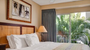 A hotel room with a bed, a nightstand, a lamp, and a view of palm trees through a window.