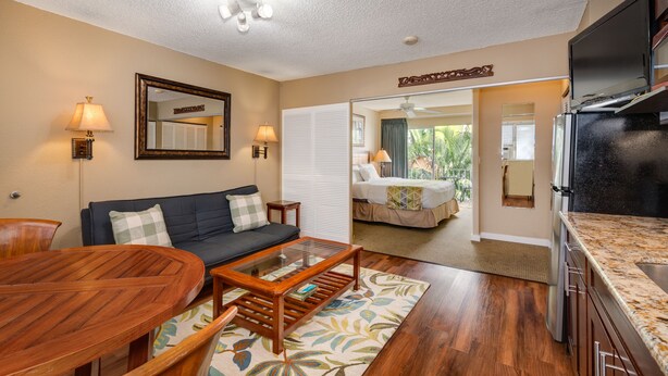 A compact living space with a sofa, coffee table, and dining table. A kitchen area with a refrigerator and stove is visible. The room has a ceiling fan and a large mirror on the wall.