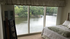 Un dormitorio con una ventana grande que da a un lago, una cama con colcha verde y un mueble de televisión con televisor de pantalla plana.