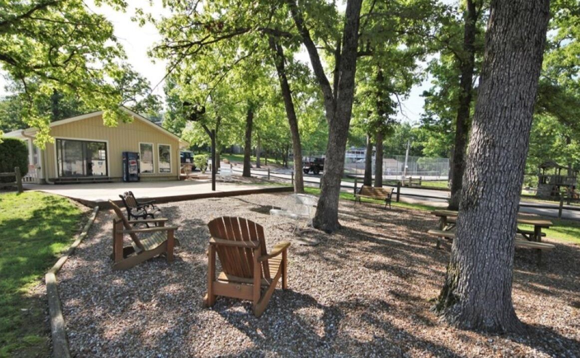 An outdoor seating area with Adirondack chairs and a wooden bench, surrounded by trees and a gravel path.