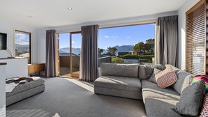 Una sala de estar moderna con un sofá gris, una pequeña mesa de centro y un televisor de pantalla plana. Hay ventanas grandes con cortinas, que ofrecen vistas a montañas y una piscina.