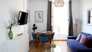 A modern living room with a blue sofa, a glass coffee table, a dining area with a wooden table and chairs, a TV mounted on the wall, and a window with curtains. - Tetuan 3I - Two Bedroom Apartment, Sleeps 4 (Sevilla)