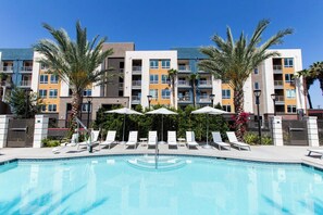 A swimming pool with lounge chairs and umbrellas, surrounded by palm trees and a multi-story building with balconies.