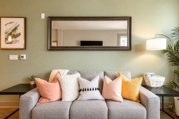 A grey sofa with multiple cushions, a large mirror above it, and a small table with a lamp and a basket.