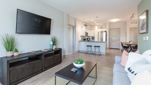 A modern living room with a flat-screen TV, dark wood entertainment unit, and a coffee table with a plant. A kitchen is visible in the background with a dining area.
