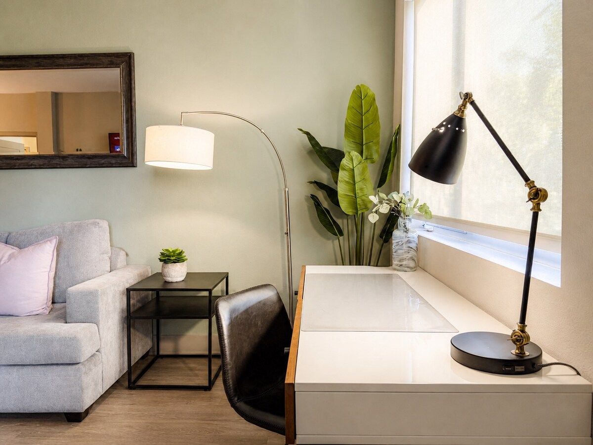 A modern room with a grey sofa, a small table with a plant, a desk with a lamp, and a large window.