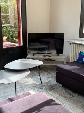 A modern living room with a flat-screen TV, a small white table, a purple sofa, and a view of greenery outside.