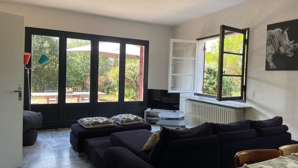 A living room with dark sofas, a TV, and a large window with a view of greenery. - Great House Montalivet (Vendays-Montalivet)