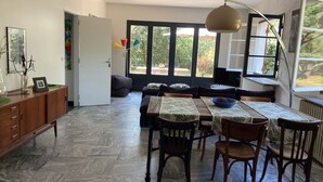 A spacious living area with a dining table, chairs, and a sofa. There is a large window providing a view of the outdoors. - Great House Montalivet (Vendays-Montalivet)