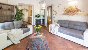 Un salón con dos sofás grises, una mesa de centro con libros y un jarrón de flores amarillas, una alfombra estampada y una pared con un cuadro floral.