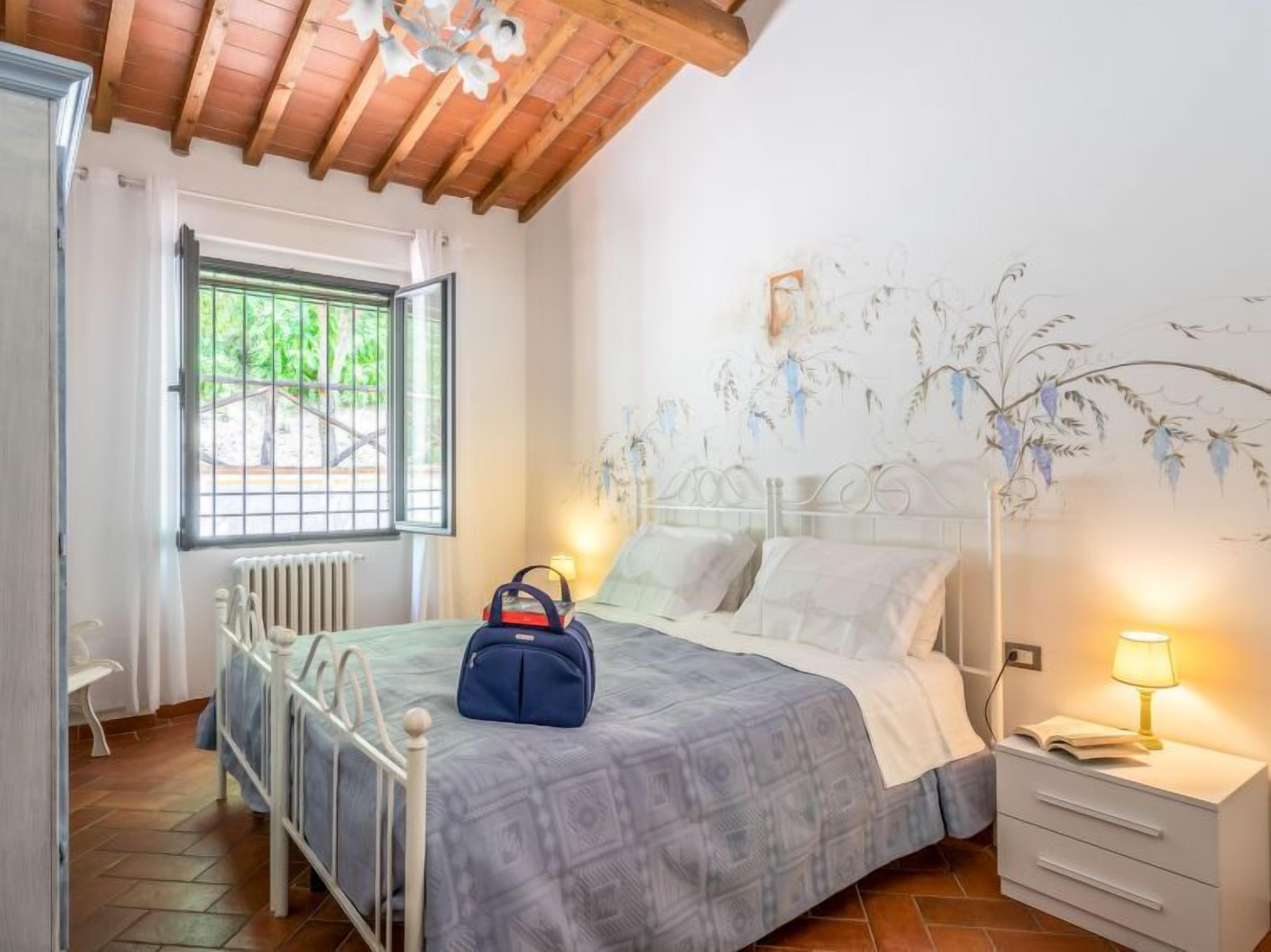 A bedroom with a bed, bedside table, lamp, window, radiator, and a blue bag on the bed.
