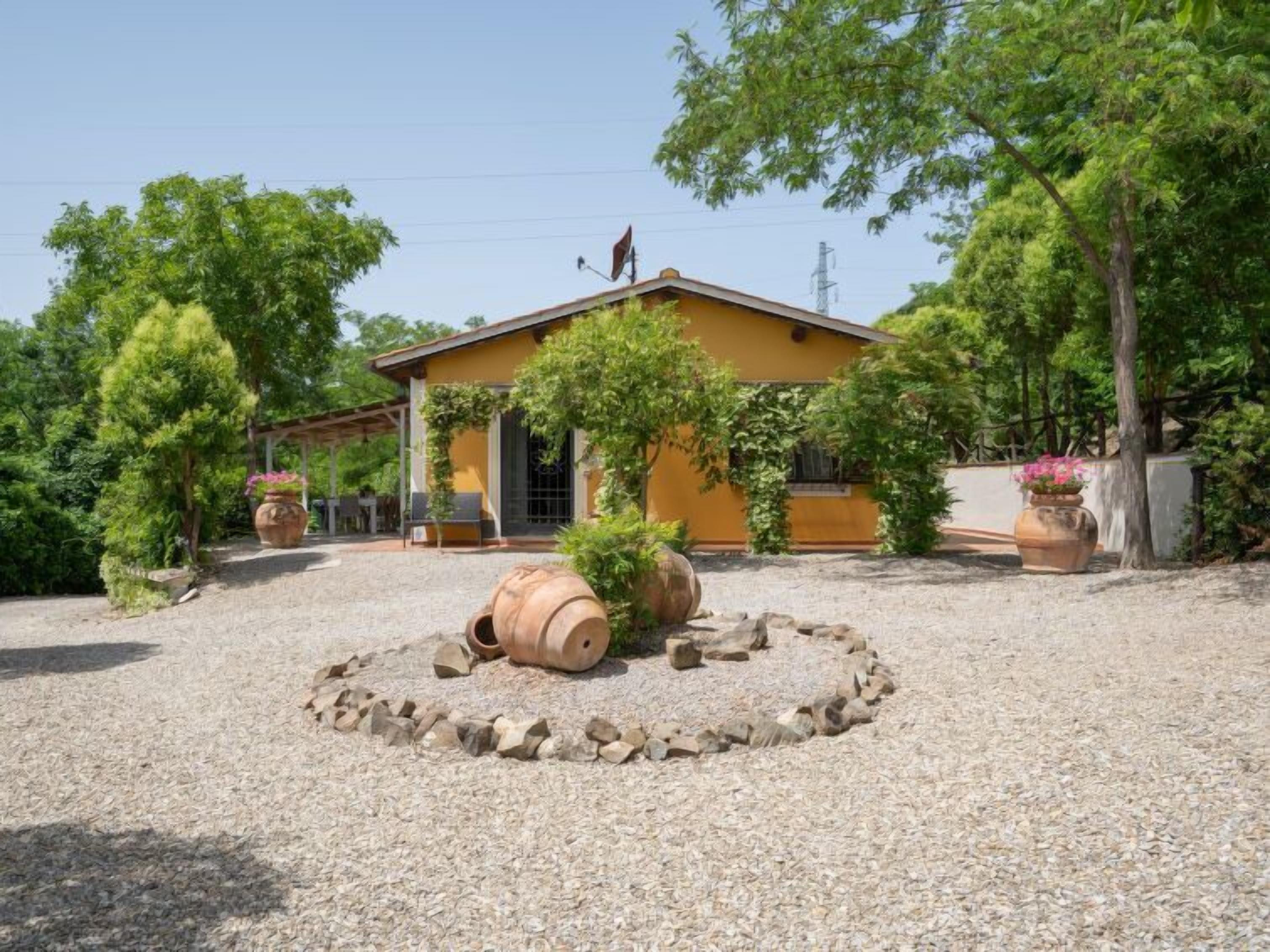 A yellow building with a gravel driveway, surrounded by trees and potted plants.