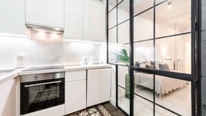 A modern kitchen with white cabinets, a built-in oven, and a large window overlooking a bedroom with a bed and a plant. - Genteel Home Velazquez II (Madrid)