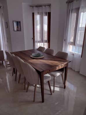 A dining area with a wooden table and six chairs, a small side table, and a window with sheer curtains.