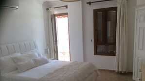 Een slaapkamer met een groot bed, een raam met gordijnen, een deur en een klein tafeltje met een lamp.