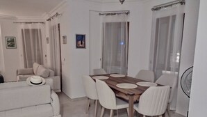 Een zithoek met een houten tafel en witte stoelen, een witte bank met een hoed erop, en een ventilator aan de muur.