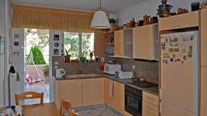 A kitchen with wooden cabinets, a dining table, chairs, a refrigerator, and a window with a view of greenery outside. - 4-rooms 145m3  ap. Seaside Athens: Glifada (Glifada)