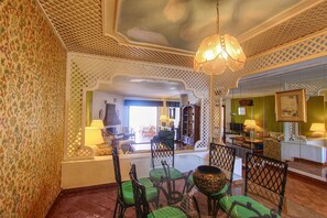 A room with a patterned wall, a chandelier, green chairs, a glass table, and a view into another room with a sofa and lamp.