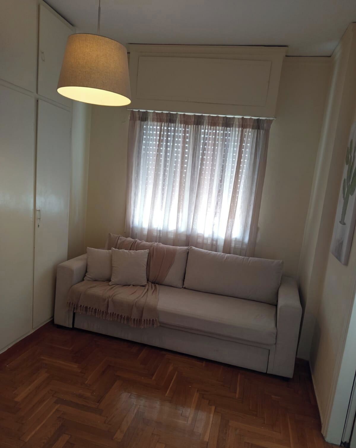 A room with a sofa, a hanging lamp, a window with sheer curtains, and a wooden floor.