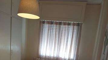A room with a sofa, a hanging lamp, a window with sheer curtains, and a wooden floor.