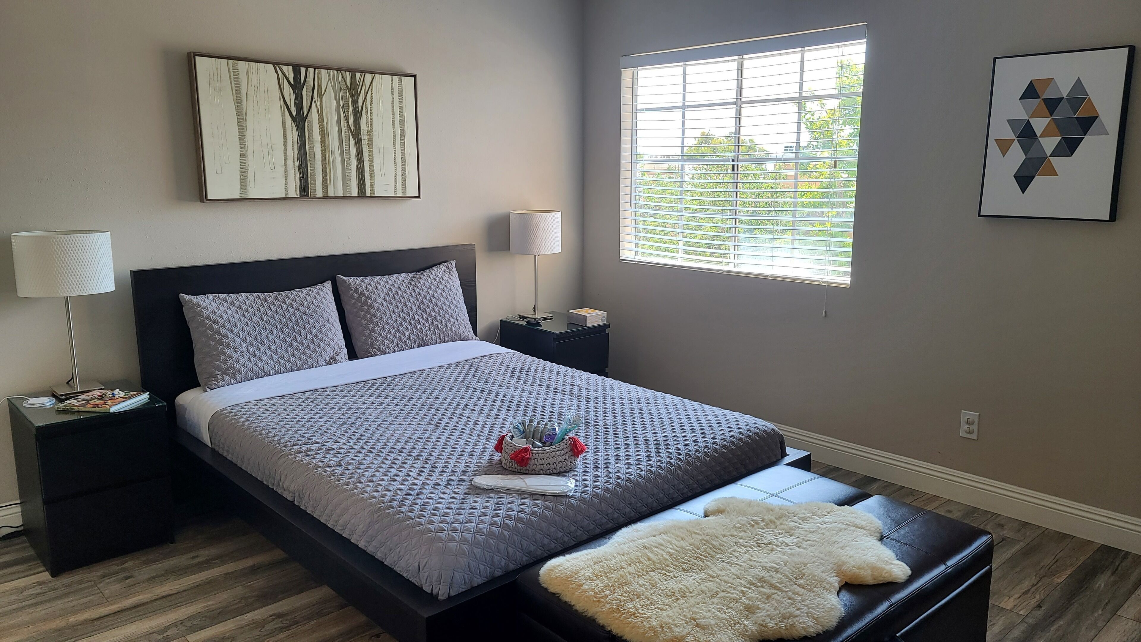 A neatly arranged bedroom with a bed, bedside tables, a window with blinds, and artwork on the walls.