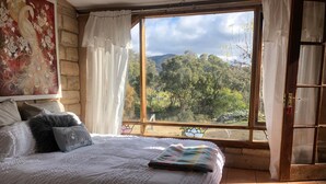 A bedroom with a large bed, a view of a forest through a window, and a painting on the wall. - Hilltop Hideaway, Firepit, Starry Nights & Privacy (Mullamuddy)