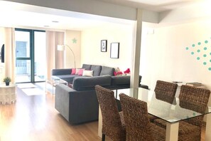 Una sala moderna con un sofá, una mesa de comedor de vidrio y sillas de mimbre. Hay un balcón con vistas al exterior.