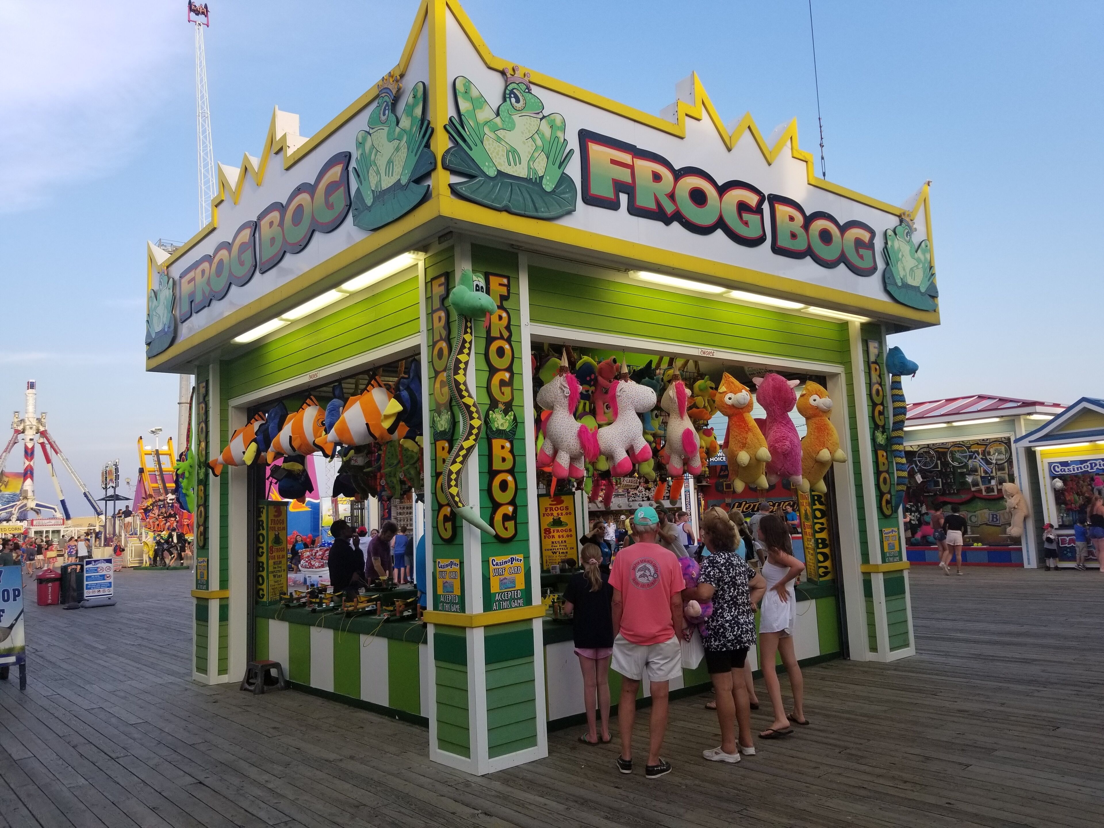 En färgstark arkadspelsautomat med namnet ”Frog Bog”, med mjuka leksaker hängande ovanför och en skylt med texten ”Casino Place” synlig i bakgrunden.