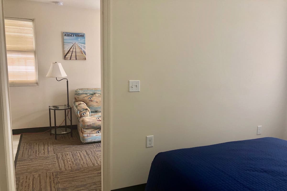 A room with a bed, a lamp, a chair, and a painting of a pier.