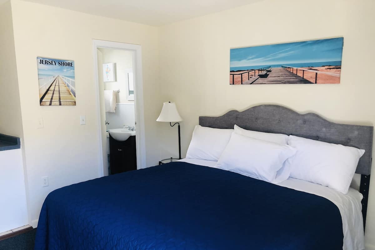 A bedroom with a large bed, a nightstand, a lamp, a painting of a beach, and a poster of a pier.